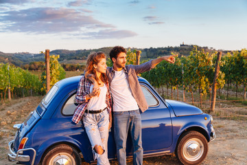 A loving couple watch the sunset, standing leaning against an old blue car in Tuscany, Italy. Around them, rows of vines and the classic hills. The young man shows to his girlfriend sunset