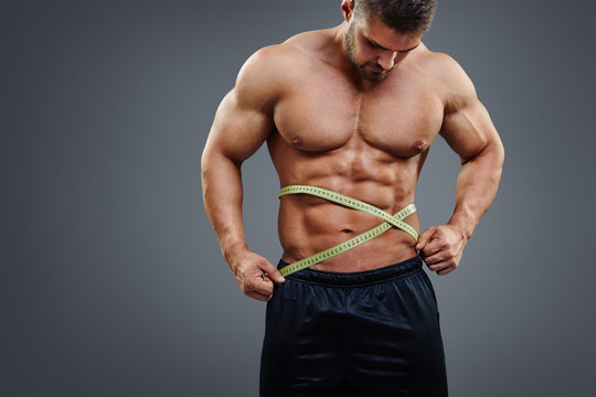 Fitness male model measuring his waist with tape measure isolated over gray background. Gain in abdominal muscles concept.