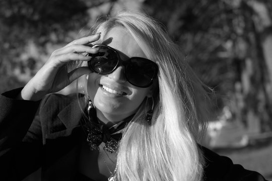 Portrait of a young beautiful woman with long blonde hair,red nails and black glasses in park. Photographed was taken on a nice sunny day. Nature background.Black and white photo.