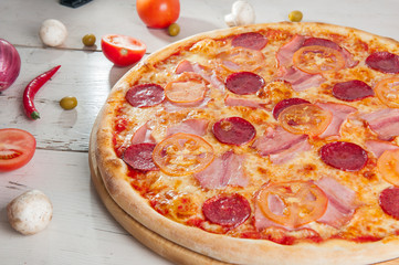  Close up Delicious Meat Pizza with Salami, Bacon and Tomato Slices on a cutting board on white wooden background with different colourful vegetables top view