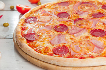  Close up Delicious Meat Pizza with Salami, Bacon and Tomato Slices on a cutting board on white wooden background with different colourful vegetables top view