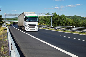 White truck passing through the electronic toll gates on the asphalt highway in a wooded landscape. Bridge and forested mountains in the background. White clouds in the blue sky.