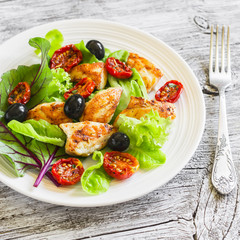 Fresh salad with chicken breast, sun-dried tomatoes, green salad and olives on a white plate on a wooden surface. Healthy food