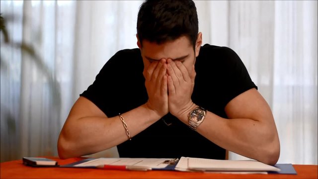 Tired, exhausted young man yawning while working or studying at home sitting at table with paper sheets in front of him
