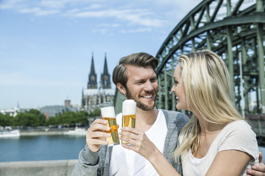 Germany, Cologne, happy young couple toasting with Koelsch glasses in front of Rhine River