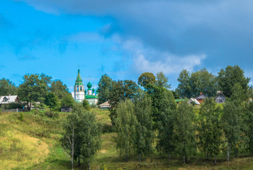 Fototapeta premium dark skies rolling in over community of Russian homes and white church with bell tower with green roof and domes 