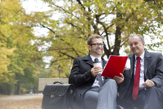 Two happy businessmen with document and coffee to go on park bench