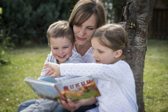 Mother and her two children sitting on a meadow reading a picture book