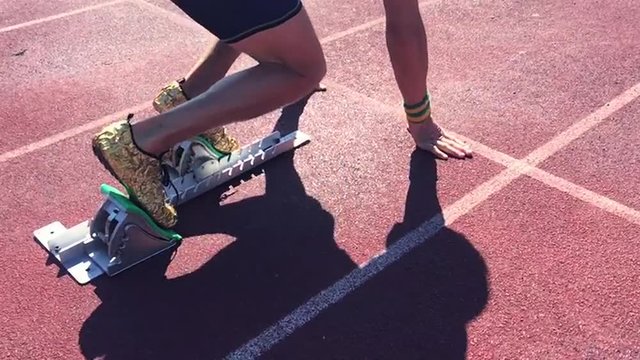 Athlete in gold shoes sprinting in slow motion from the starting blocks over the starting line of a race on a red running track 