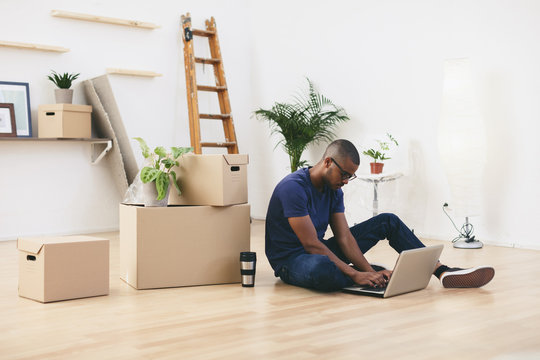 Young man sitting beside cardboard boxes in his new flat using laptop