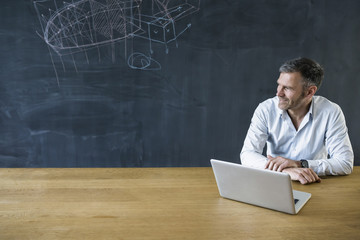 Smiling mature man with laptop at blackboard