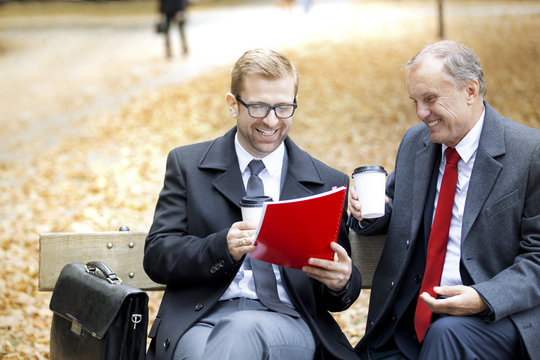 Two happy businessmen with document and coffee to go on park bench