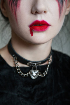 Young beautiful gothic woman with white skin and red lips with bloody drops wearing black collar with spikes. Red smokey eyes. Halloween makeup.