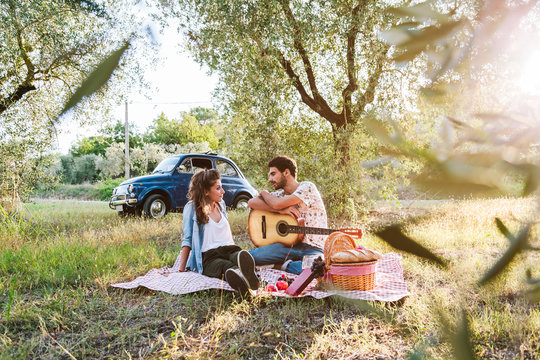 On a beautiful sunny day, a couple of young lovers, makes picnic on grass among olive groves in Tuscany, Italy. Man leans on guitar while talking with his girlfriend. Behind them a blue vintage car