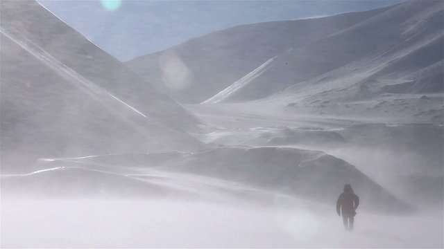 A lone traveller is walking through a snowstorm on a background of snow-capped mountains.
