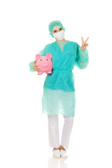 Female surgeon doctor with piggy bank shows victory sign
