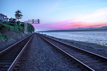 Fototapeta na wymiar Train Tracks Vanishing into Distance on Ocean Beach at Sunset with Pink and Purple Mountain Range in Background