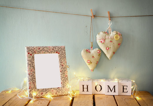 image of blank frame next to wooden blocks with word home, and hanging fabric hearts on rope, garland lights in front of wooden background. retro filtered image