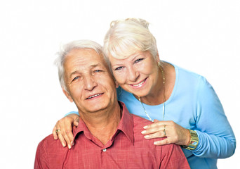  Seniorin  steht hinter ihrem sitzenden Mann und umarmt ihn von hinten, beide lächeln in die Kamera