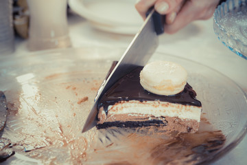 Female hand with a knife slicing chocolate-vanilla mousse cake with Glsaz of chocolate and white macaroon on it. 
