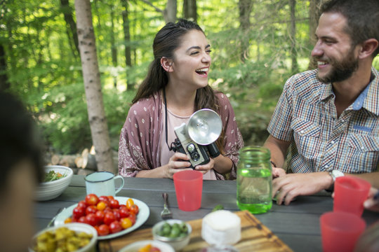 A group of people seated at a meal around a table outdoors. A couple side by side, one holding a camera with separate metal flash unit. 