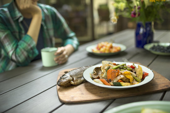 A person seated at a table outdoors. A cooked fresh fish and dish of vegetables. 
