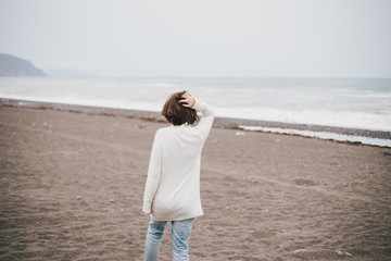 Beautiful young woman wearing white sweater and blue jeans walking on a lonely beach in a cold windy weather