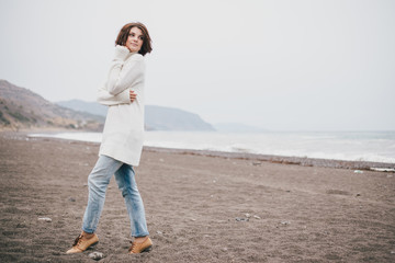 Beautiful young woman wearing white sweater and blue jeans walking on a lonely beach in a cold windy weather