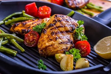 Grilled chicken breast in different variations with cherry tomatoes, green French beans, garlic, herbs, cut lemon on a wooden board or teflon pan. Traditional cuisine. Grill kitchen. 