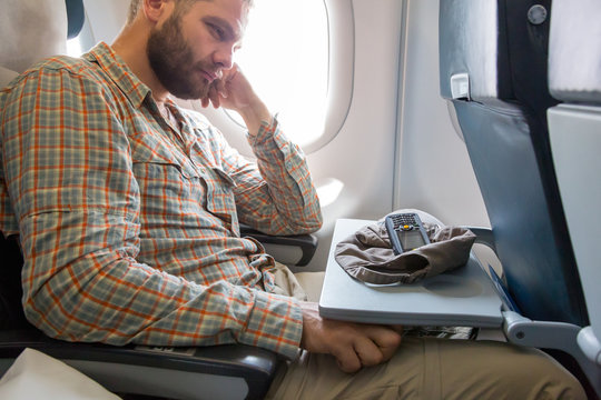 Tired Traveler at Airplane Passenger Chair Bearded Young Man Casual Clothing Sitting next to Aircraft Window with Cap and Telephone Dropped on Unfolded Table
