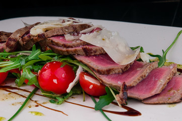 Medium rare steak served on a plate with cherry tomatoes and arugula. On top of it is grated parmesan cheese.