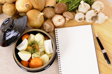 Casserole pot or stockpot crock with vegetables and herbs on kitchen worktop with blank recipe book or cookbook making preparing vegetable soup or stew photo 