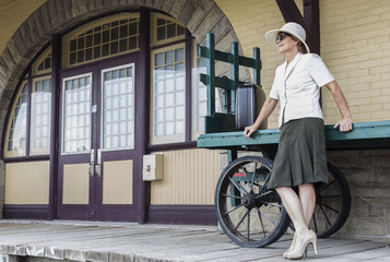 Fototapeta na wymiar horizontal image of a woman wearing a dress and a big hat standing by an old luggage cart with her suitcase sitting on it by the doors of an old train station.