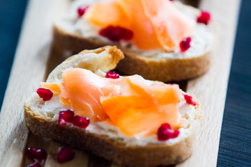 Salmon bruschetta with white cheese and pomegranates on wooden kitchen board over dark wooden background
