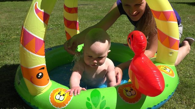 Beautiful mother refresh cute infant baby in kiddie pool water on hot day. Zoom out shot. 4K
