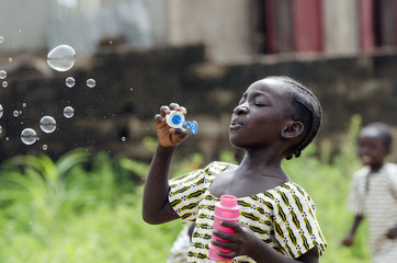 African Girl Playing with Soap Bubbles Outdoors in Bamako, Mali. Black young beautiful girl having fun outdoors blowing soap bubbles. Happiness symbol. Elementary student happy outside her school. - obrazy, fototapety, plakaty
