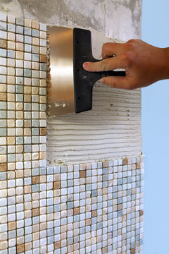 Repairs in the apartment: installing mosaic tile on wall. / A worker covers the wall with adhesive using a notched trowel.