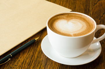 A cup of coffee with heart shape, pen and blank book on wood bac