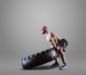 Athletic young man lifting a tire on gray studio background
