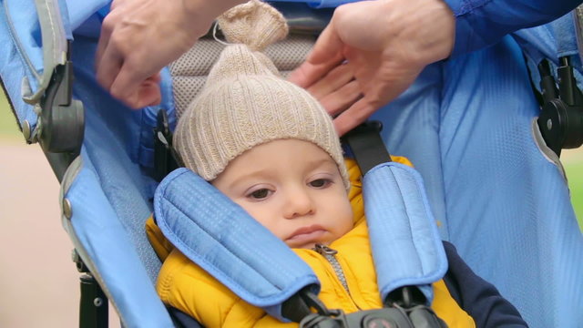 Sad baby boy sitting in buggy stroller, his mother pulling down knit hat over his ears 
