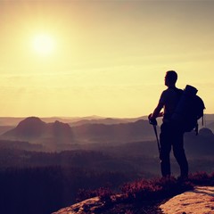Fototapeta premium Tall backpacker with poles in hand. Sunny daybreak in mountains. Hiker with big backpack on rocky view point above misty valley.