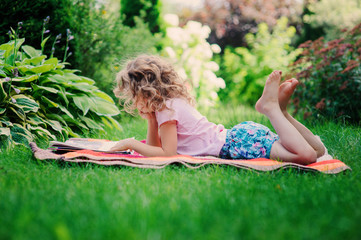 child girl reading on summer vacation in sunny garden on blanket