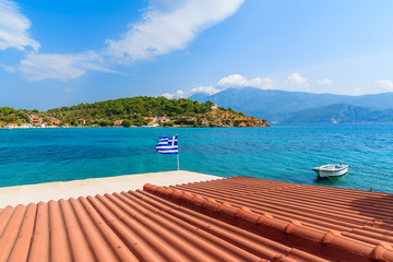 Obraz premium Orange tile roof of a house with Greek flag waving against blue sea background on coast of Samos island, Greece