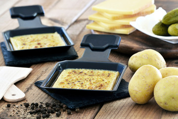 Raclette - Raclettekäse, gebraten / In Pfännchen mit Kartoffeln, Gewürzgurken und Essigzwiebeln - Schweizer Spezialität / Nahaufnahme - Makro / Rustikal auf Holz