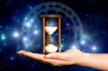 female hand holding an hourglass over blue mystical background with stars and astrology chart