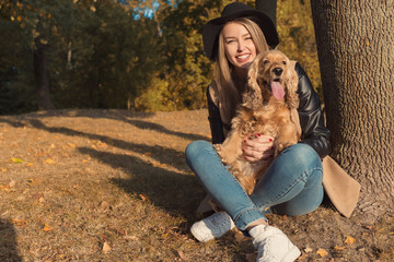 Beautiful cute happy girl in a black hat playing with her dog in a park in autumn another sunny day