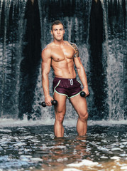 Muscular man lifting dumbbell on waterfall
