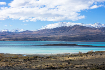 Lake Tekapo in beautiful turquoise blue, that is caused by glacial flour, fine rock particles from the glaciers. The lake is dominant among brown dry grass, and dark-toned color of mountain ridges.