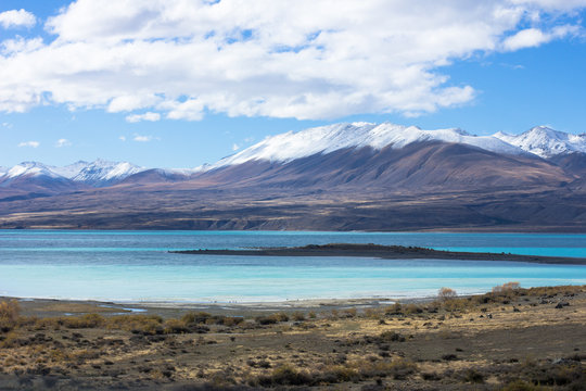 Lake Tekapo in beautiful turquoise blue, that is caused by glacial flour, fine rock particles from the glaciers. The lake is dominant among brown dry grass, and dark-toned color of mountain ridges.
