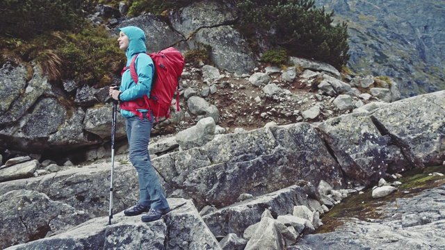 Hiker woman standing with backpack on the edge of the cliff admiring mountain lake view landscape. Stabilized, 4K Ultra HD. Epic Steadicam hiking in stormy wind, lifestyle. Misty Mountains Series. 